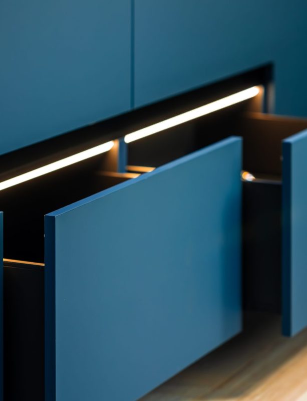 A linear LED strip highlights a run of half open drawers, adding rhythm across matte blue fronts for a sleek, modern kitchen.